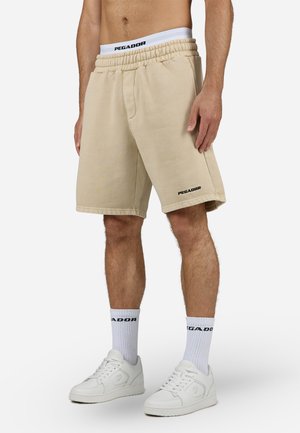 Beiga bomullsshorts med elastisk midja, med sidofickor och en svart "PEGADOR"-logotyp på vänster ben. Bärs med vita sneakers.