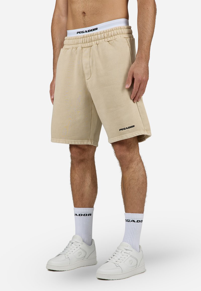 Pantalones cortos de algodón beige con cintura elástica, con bolsillos laterales y un logo negro "PEGADOR" en la pierna izquierda. Llevados con zapatillas blancas.