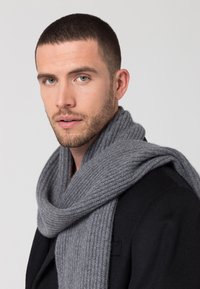 Sciarpa grigia lavorata a maglia in morbida lana, con una texture a coste. Indossata avvolta attorno al collo, completa un cappotto nero.