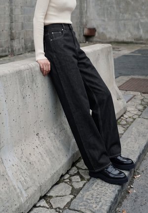 Personne portant un jean noir à jambes larges, des chaussures noires épaisses, et un haut à manches longues côtelé crème, appuyée contre une barrière en béton à l'extérieur.