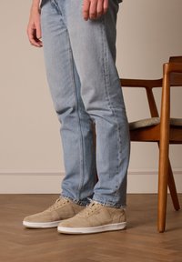 Sneakers in suede beige con suole in gomma bianca, abbinati a jeans azzurri chiari. L'ambientazione presenta un caldo pavimento in legno e una sedia in legno.