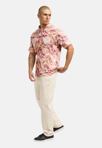 Kurzärmliges Hemd in Pink und Creme mit Muster und dem Text "Culture", kombiniert mit beigen Hosen und schwarzen Sneakers, Model posiert seitlich.