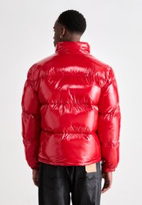 Red puffer jacket featuring a glossy finish, high collar, and quilted design. The sleeves have elastic cuffs and a tag on the back.