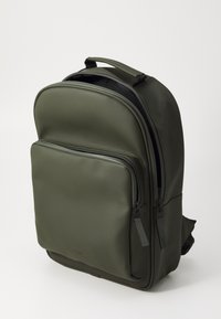 Un sac à dos vert olive, fabriqué en matériau lisse, possède un compartiment principal zippé, une poche avant, et des zips et des accents noirs.