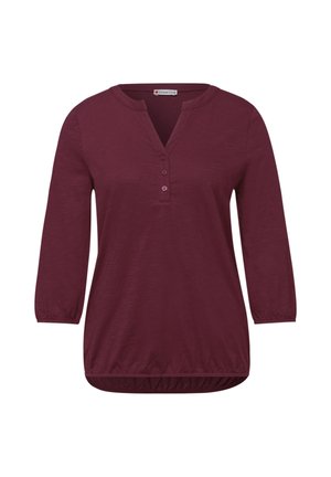 Luumuinen pitkähiha-paita, jossa on kolme nappia, pyöreä kaula-aukko uralla sekä joustavat hihansuut ja helma.