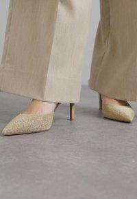Gold glitter high-heeled shoes with a pointed toe design, featuring a smooth texture and slender heel, paired with beige wide-leg trousers.
