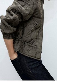 Quilted olivengrøn bomberjakke med elastiske manchetter, sidelommer og tekstureret stof, parret med mørk denim jeans.