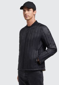 Black padded jacket with vertical quilted pattern, zip closure, and ribbed collar, paired with a black cap and dark jeans.
