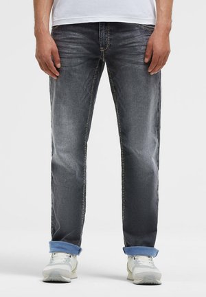 Mann trägt ausgewaschene dunkelgraue Jeans mit aufgekrempelten Säumen und weißen Sneakers, steht vor einem schlichten hellen Hintergrund.