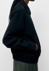 Giacca nera strutturata con collo e orlo elastico. Presenta maniche lunghe e tasche laterali. Realizzata in tessuto liscio e leggero.