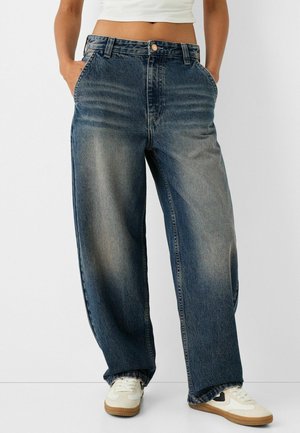 Kvinde iført løstsiddende, falmede blå jeans og hvide sneakers, stående med hænderne i lommerne mod en ensfarvet baggrund.
