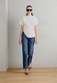 Top blanco de manga corta con diseño asimétrico, combinado con jeans rectos azules y tacones de print de leopardo, frente a una pared neutral.