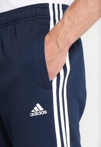 Main dans la poche d'un pantalon Adidas bleu marine avec trois bandes blanches et logo, taille élastique visible.