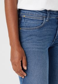 Vaqueros de mezclilla azul con corte slim, con herrajes en tono dorado, bolsillos y una textura suave. La cinturilla se sitúa por encima de las caderas.