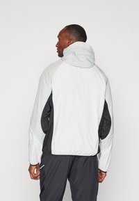 Veste légère grise avec des accents noirs, dotée d'une capuche, de coutures réfléchissantes et d'une finition texturée. Convient pour une tenue sportive.