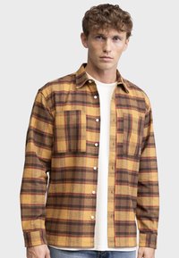 Cămașă din flanel în carouri galbene și maro, cu mâneci lungi, închidere cu nasturi, două buzunare la piept și un tricou alb cu gât rotund dedesubt.