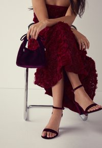 Šaty s karmínovým růžovým vzorem, sametová bordó kabelka s zakřiveným vrškem a stuhovými držadly a sandály s tenkým podpatkem a řemínky v barvě bordó.
