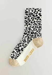 Schwarz-weiße Leopardenmuster-Socke mit goldfarbener Zehenakzentuierung und goldfarbener Logobeschriftung an der Seite. Das Material wirkt gestrickt und weich.