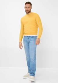 Gelber Rundhalsausschnitt-Pullover aus leicht strukturiertem Stoff, getragen über einem weißen T-Shirt, kombiniert mit hellblauen Jeans und beigen Sneakers.