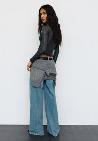 Frau mit langen, gewellten Haaren, die ein dunkles Spitzentop, weite Jeans und einen grauen Pullover, der um ihre Taille gebunden ist, trägt und vor einer schlichten weißen Wand steht.