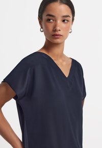 Marinblå V-ringad topp i lätt material, med korta ärmar, en avslappnad passform och subtila sidoklinningar för ökad rörelsefrihet.