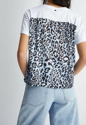 Vrouw draagt een wit shirt met korte mouwen met een blauwe en zwarte luipaardprint op het achterpaneel en lichtblauwe high-waisted spijkerbroek, gezien van achteren.