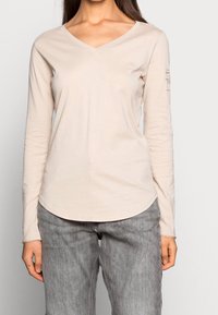 Långärmad beige T-shirt med V-ringning och rundad nederkant. Har en liten logotyp på bröstet och tryckt text på ärmen. Mjuk bomullsmaterial.