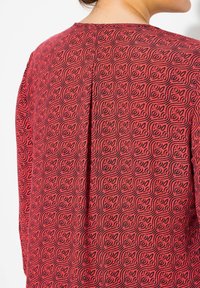 Rotes Top mit Muster, das ein strukturiertes Design mit Wellen und Kurven in Schwarz zeigt. Verfügt über einen schlichten Rückenausschnitt und Faltendetails.