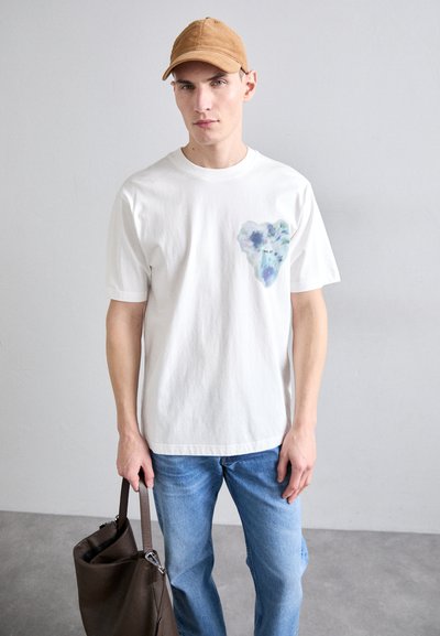 Camiseta blanca de algodón con un diseño de corazón azul y morado en el lado izquierdo del pecho. El modelo lleva puesta unos jeans de color azul claro y sostiene una bolsa marrón.