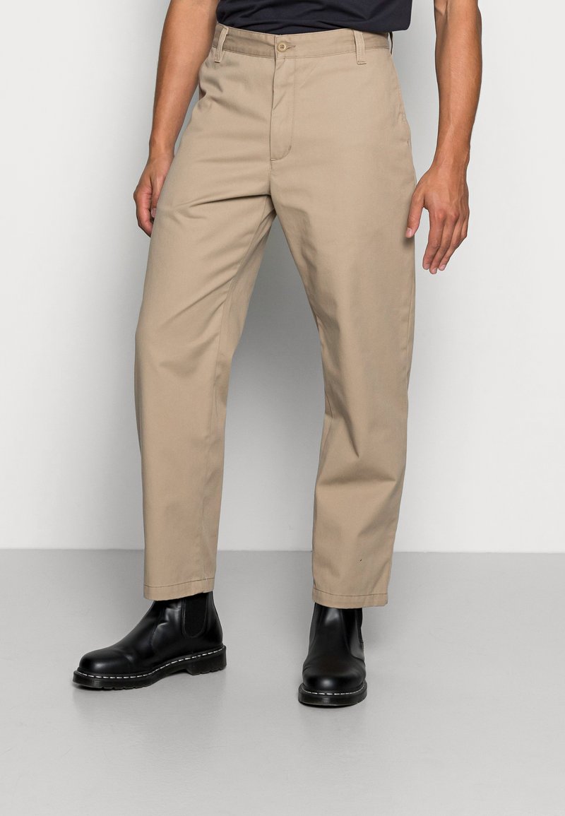 Chinos de algodão bege com pernas retas, bolsos frontais e fecho com botão, emparelhados com botas de tornozelo pretas com sola texturizada.