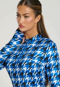 Langsleeve top gemaakt van rekbare stof, met een blauw, zwart en wit houndstoothpatroon, een half-rits kraag en een nauwsluitend ontwerp.