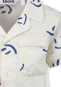 Kortärmad krämfärgad skjorta med blått abstrakt leende ansiktsmönster. Har krage, knappstängning och bröstficka. Mjuk tyg.