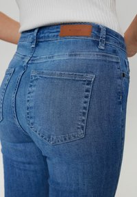 Zilā denim jostas ar vidēju vidukļa piegriezumu, ar ādas brenda uzlīmi, klasisko piecu kabatu dizainu un mīkstu, nedaudz izbalējušu tekstūru.