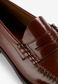 Mocasín de cuero marrón con un acabado brillante, que presenta costuras detalladas y un acento trenzado en la parte superior. Forma redondeada, textura suave.