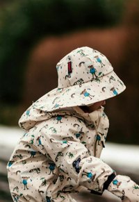 Wasserfeste Jacke und Sonnenhut aus leicht beigem Stoff, verziert mit einem verspielten Druck verschiedener Tiere mit Accessoires und Regentropfen.