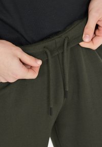 Olijfgroene sweatpants met een elastische tailleband en een trekkoord. De stof lijkt zacht en heeft een gladde textuur, zonder zichtbare patronen.