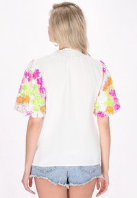 Hvid tekstureret bluse med farverige blomster-sequin detaljer på puffærmerne. Bagfra ses en afslappet pasform og korte, flossede denimshorts.