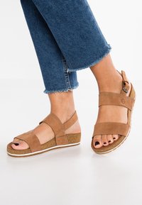 Tan kile sandaler laget av myk semsket skinn, med korslagte stropper og justerbar ankelstropp. Rund tå med en teksturert korksåle.