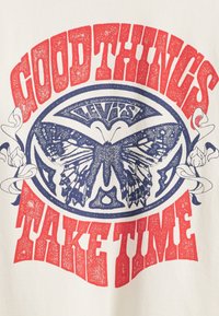 Weißes T-Shirt mit einem Schmetterlingsmotiv in Blau und Rot sowie dem Text "GUTE DINGE BRAUCHEN ZEIT" in einer fetten, abgenutzten Schrift.