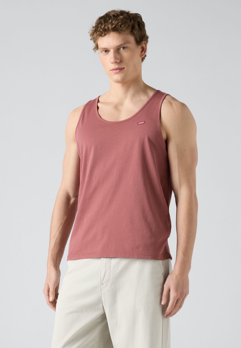 Junger Mann mit lockigem Haar, der ein ärmelloses Tanktop in staubigem Rosa und hellbeige Hose trägt und vor einem schlichten Hintergrund steht.