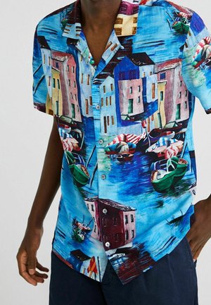 Hemd mit lebendigem Küstendorf-Motiv, das bunte Häuser, Boote auf dem Wasser und einen strahlend blauen Himmel zeigt, kombiniert mit dunklen Shorts.