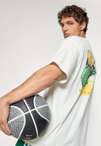 Hvid t-shirt med korte ærmer og grafisk design på bagsiden, holder en struktureret sort og grå basketball med hvide accenter.