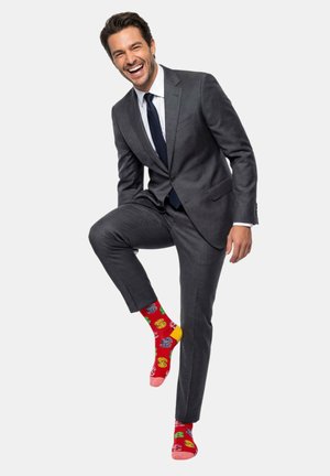 Mies tummanharmaassa puvussa ja tummansinisessä solmiossa nauraa, nostaen yhtä jalkaa paljastaakseen kirkkaanpunaiset sukat, joissa on värikkäitä abstrakteja kuvioita.