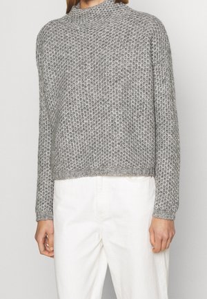 Kobieta ubrana w szary, gruby sweter z dzianiny z długimi rękawami, zestawiony z białymi, wysokim stanem spodniami, obciętymi na wysokości talii.