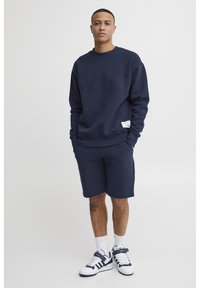 Marineblå hettegenser og matchende shorts laget av myk stoff. Har rund hals og en liten etikett på nederste venstre del av hettegenseren.