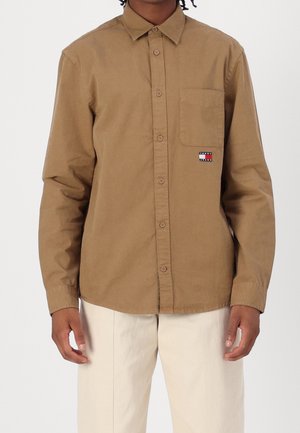 Homme portant une chemise marron à manches longues boutonnée avec une poche sur la poitrine et un pantalon beige, debout devant un fond uni.