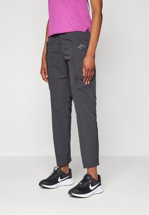 Personne portant un pantalon noir fuselé, des baskets Nike noires et blanches, et un haut à manches courtes rose, debout devant un fond uni.