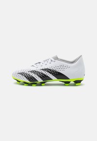 Scarpino da calcio bianco con strisce diagonali nere, parte superiore perforata, linguetta grigia e suola verde brillante per una presa ottimale su superfici erbose.