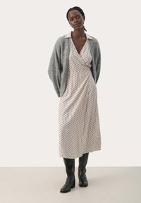 Siva cardigan preko bež obleke s pikami in ovratnikom, srednje dolžine. Model nosi črne škornje do kolen. Gladek material, sproščen kroj.