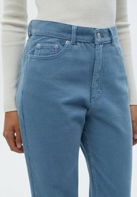 Pantalon en velours côtelé bleu clair avec texture cannelée verticale, conception à cinq poches et éléments métalliques, associé à un haut tricoté crème.